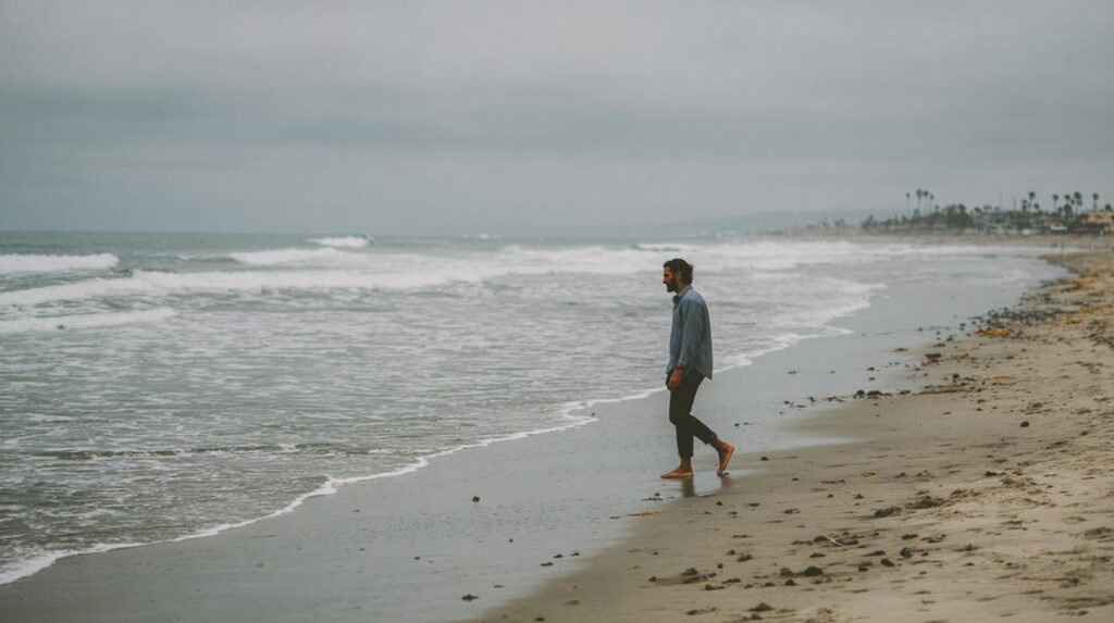 A man walks barefoot along a cloudy shoreline, reflecting quietly as if seeking therapy for feeling overwhelmed.