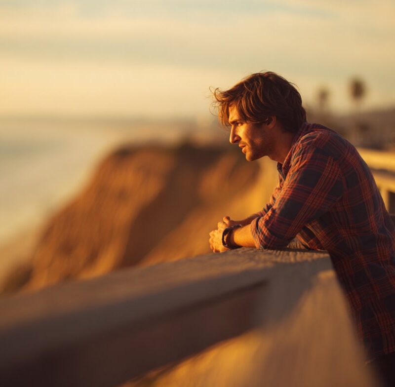 A man leaning on a coastal overlook at sunset deep in thought about his life and emotions, reflecting the question why do I feel empty.